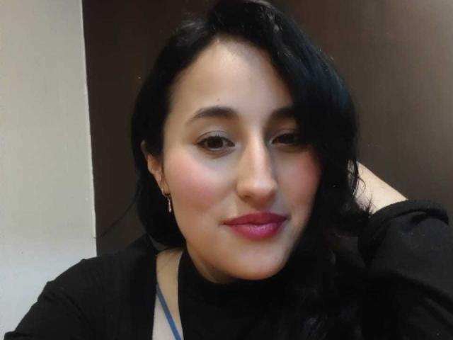 MaiGolf has dark, wavy hair, pink lipstick, and is wearing a black top with a blue lanyard, smiling softly with her right hand in her hair.