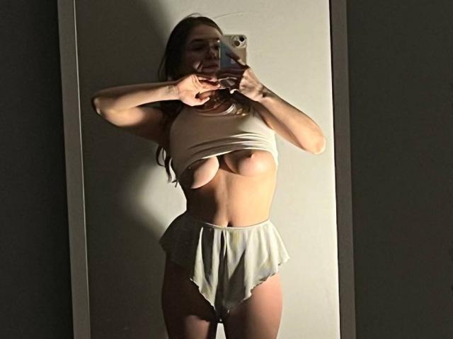 LinaStones with exposed tits in a beige crop top and high-waisted, frilled white panties, taking a mirror selfie, holding her phone with a peace sign.