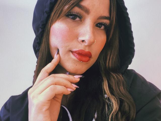 Sara Duncan tiene lápiz labial rojo, un piercing en la nariz y cabello largo y castaño, lleva una capucha negra, y tiene las uñas rosas tocando su mejilla. Está mirando a la cámara con una ligera sonrisa..