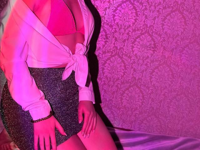 LisaAlisia with pink bra, white tied shirt, and sparkly black skirt, hands on thighs, pink and purple lighting, white patterned wallpaper, bracelet on right wrist.