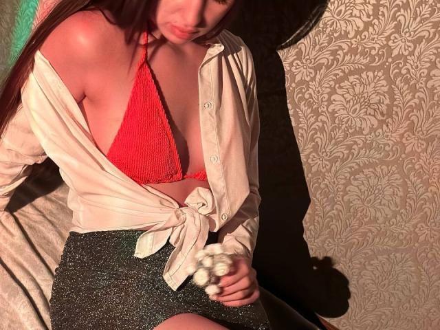 LisaAlisia with red bikini top, showing her tits, wearing a tied white shirt and sparkly black skirt, holding a fluffy toy, in dim lighting.