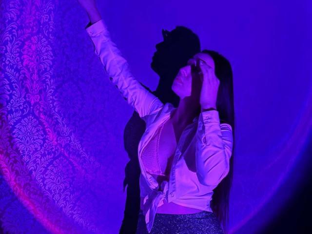 LisaAlisia with large tits, wearing a white shirt tied at the front, silver sparkly pants, hands on her head, arm raised, shadow on the wall, purple blue lighting.