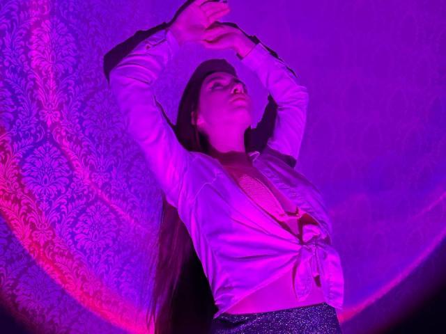 LisaAlisia with arms raised, wearing a tied-up white shirt, exposing her tits, and a sparkly skirt, under purple and pink lighting. She's looking up, with a confident expression.