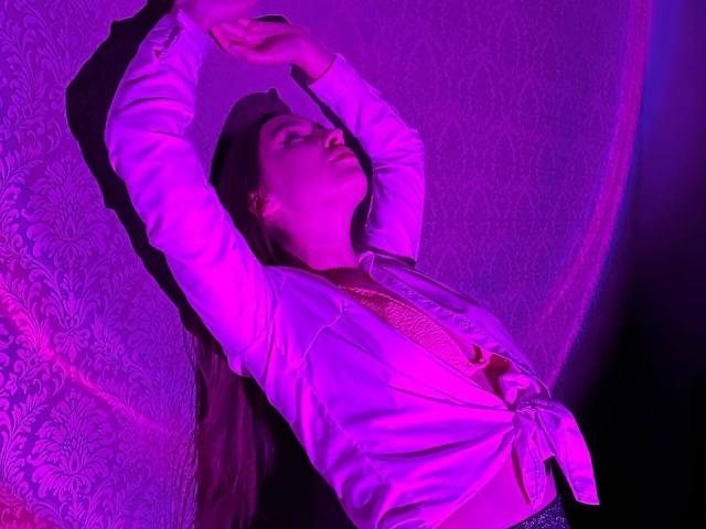 LisaAlisia with arms raised, wearing a tied-up white shirt, pink lighting, long brown hair, eyes closed, pink and purple hues, relaxed and sensual, soft shadows, moody atmosphere.