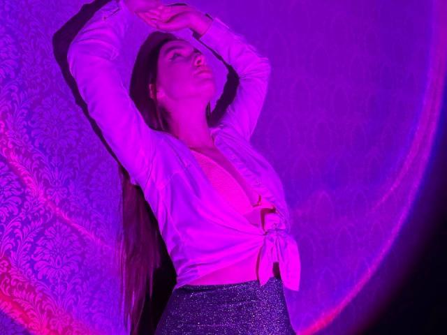 LisaAlisia with arms raised, wearing a tied-up white shirt exposing her tits, and a sparkly purple skirt, illuminated by pink and purple lighting.