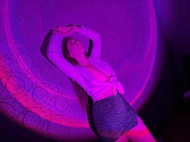 LisaAlisia with her arms above her head, wearing a tied white shirt and sparkly gray mini-skirt, under purple and pink lighting, looking sexy and confident.