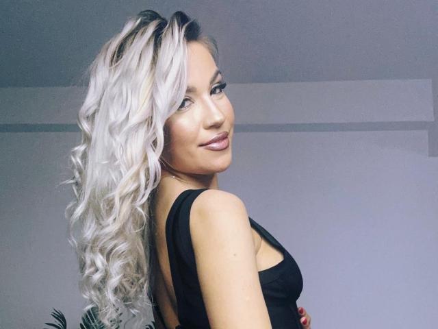 ZoeyMarie, avec de longs cheveux blonds platine bouclés, portant un haut noir, regardant par-dessus son épaule, souriant, montrant ses fesses, dans un environnement intérieur bien éclairé..