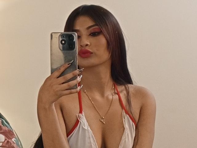 EmmaaRodriguez with long brown hair, red lipstick, and red eyeliner, holding a phone, wearing a white and red top, showing her tits, in a mirror selfie.