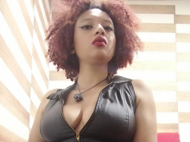 VioletaSwitch with curly red hair, wearing a black leather zip-up top, showing big tits, red lipstick, and a necklace with a black cat pendant. Low angle shot.