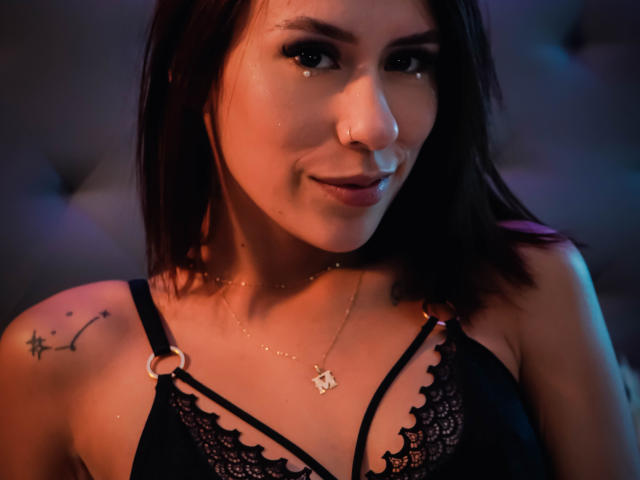 MilaKat with dark brown hair, nose ring, and black lace bra, showing her small tits and a star tattoo on her right shoulder. Soft lighting highlights her cute smile.