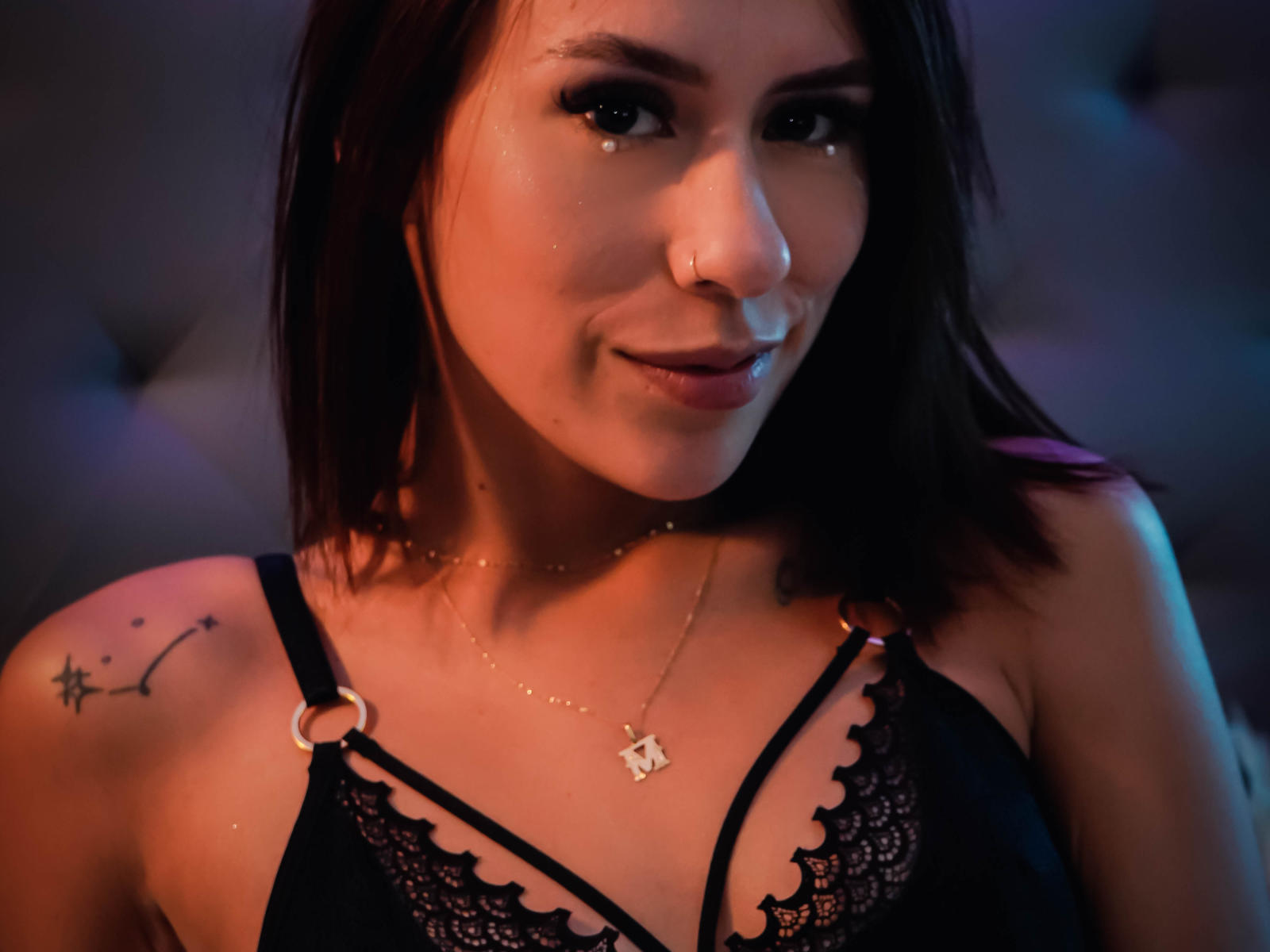 MilaKat with dark brown hair, nose ring, and black lace bra, showing her small tits and a star tattoo on her right shoulder. Soft lighting highlights her cute smile.