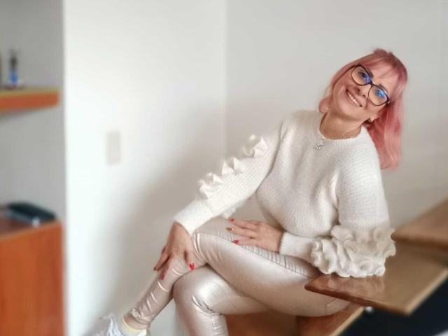 JaneTurner with pink hair, glasses, white sweater, and shiny silver pants, sitting on a wooden counter, smiling, hands on her knee, red nails, white sneakers.