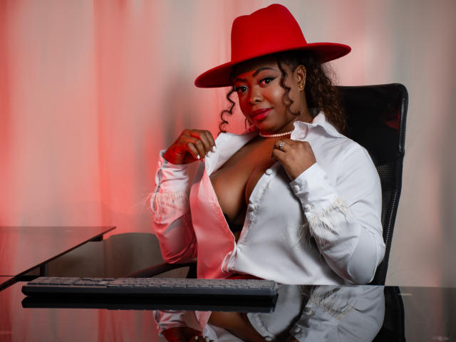 EbonnyTitsBig with curly brown hair, red hat, white shirt, and exposed large tits, sitting at a glass table with a keyboard, looking at the camera with a smile.