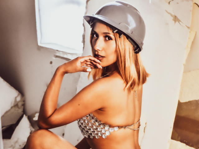 BrianaHarpers with blonde bob, wearing a shiny helmet and a sparkling bralette, looking over her shoulder with a finger to her lips, showing her bare ass.