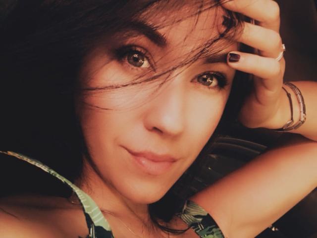 Misstyk has brown eyes, dark hair, and a slight smile. She's touching her hair with a hand showing black nails and bracelets. She's wearing a green patterned top.
