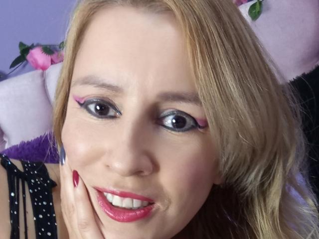 MadissonS has blonde hair, pink eyeliner, and red lipstick. She's smiling with her hand on her cheek, showing red nails. Her tits are not visible.