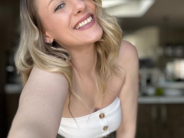 HottieAshleeyy with blonde wavy hair, smiling, wearing a white strapless top with buttons, showing her boobs, standing in a kitchen, holding a selfie.