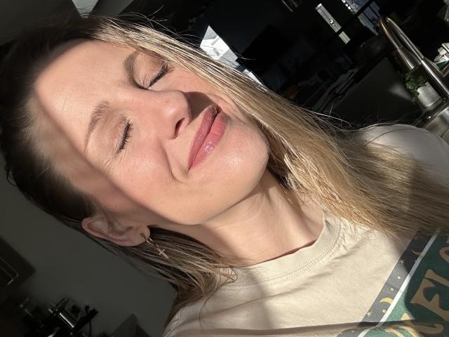 HottieAshleeyy with closed eyes, smiling, light blonde hair, white shirt, sunlight on her face, subtle tattoo on her right arm, slight sheen on her skin.