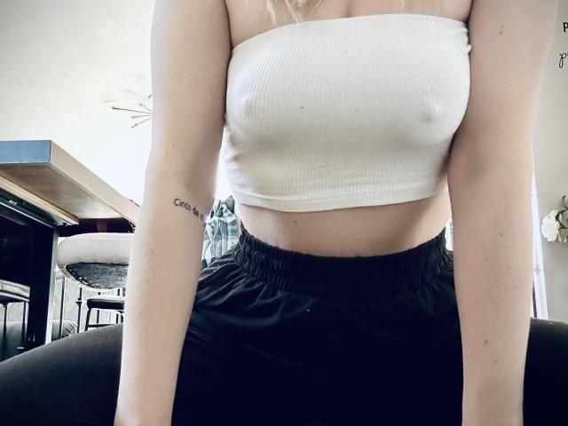 HottieAshleeyy with white tube top showing her small tits, black high-waisted pants, and "Crazy" tattoo on her right arm, standing in a softly lit room.