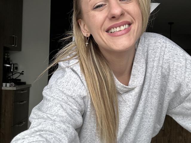 HottieAshleeyy with long blonde hair, smiling, wearing a grey sweatshirt, cross earrings, and pink lipstick, taking a selfie, showing her teeth and upper body.