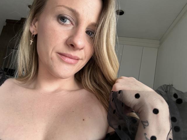 HottieAshleeyy has blonde hair, light skin, and a slight smile. She's wearing a sheer black polka dot glove, exposing her tits. She's got a cross earring.