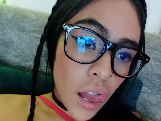 StellaBeckker with black braids, thick black glasses, and a tongue piercing, looking into a camera with her tongue out, wearing a yellow top with a red strap.