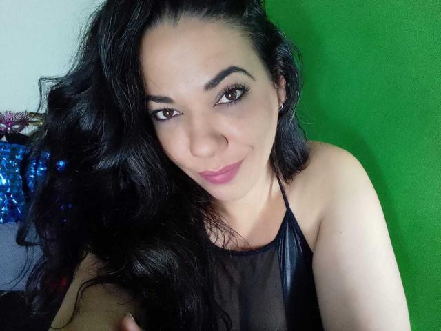 YolittS with long black hair, brown eyes, and pink lipstick, wearing a sheer black top, showing her tits, smiling at the camera against a green background.