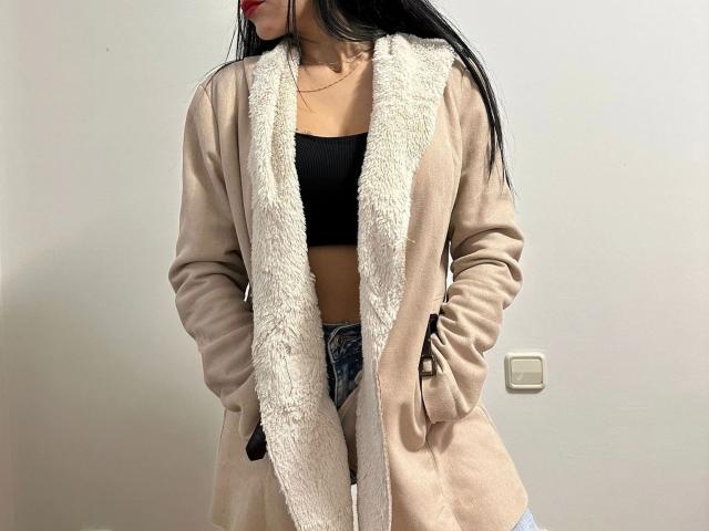 De prinses - aantrekkelijk met lang zwart haar, rode lippenstift, een beige jas met witte bontrand, een zwarte sportbeha, en met haar handen in haar zakken, draagt blauwe jeans..