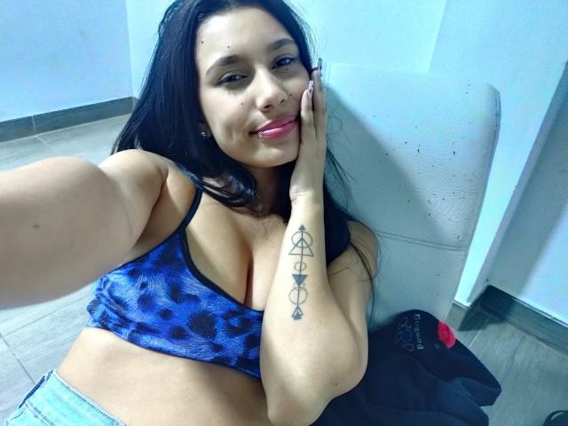 KataleyaMansson, with long black hair, pink lipstick, and blue leopard print top, showing off her tits and tattoo on her right arm, smiling at the camera.
