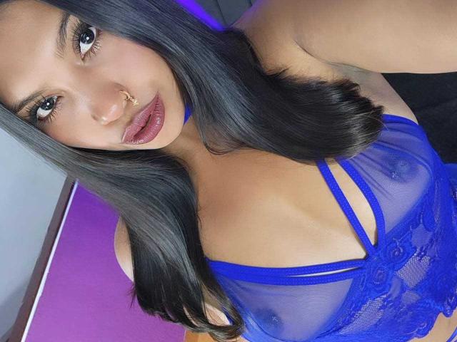 KimJohnsson a de longs cheveux noirs, des yeux bleus et un piercing au nez. Elle porte un soutien-gorge en dentelle bleu transparent, qui révèle ses seins, avec un éclairage violet en arrière-plan..