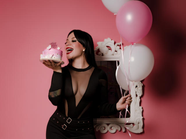 KatalinaJohnson with black mesh top, showing her tits, holding pink cake, smiling, pink and white balloons, white ornate mirror, pink wall, black belt, red lipstick.