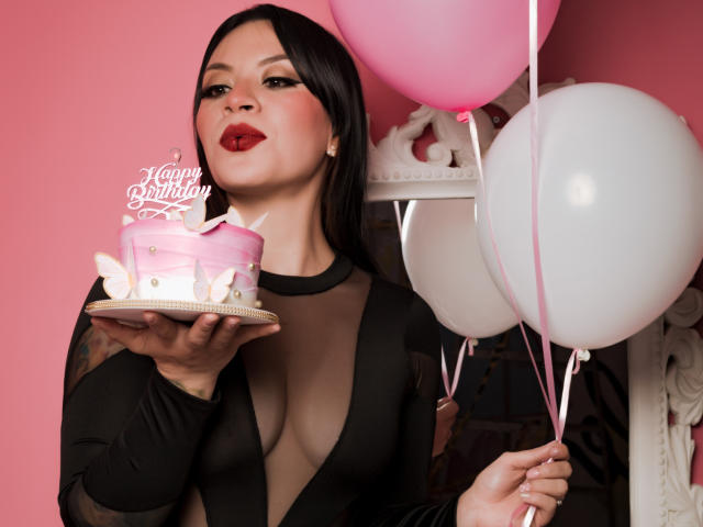 KatalinaJohnson with black hair, red lipstick, and sheer black dress, holding pink birthday cake with bunny topper, pink and white balloons in hand, looking at camera.