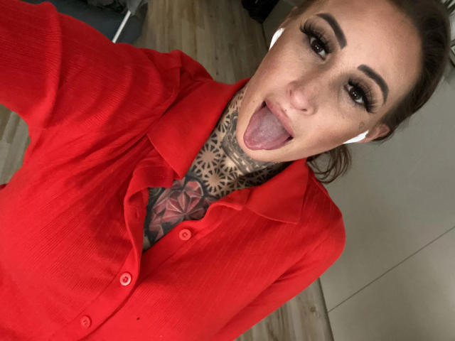 Black-Rosee-hot with a red shirt, tongue out, tattoos on her neck, wearing white earbuds, brown hair in a ponytail, heavy makeup, and a sexy, confident expression.