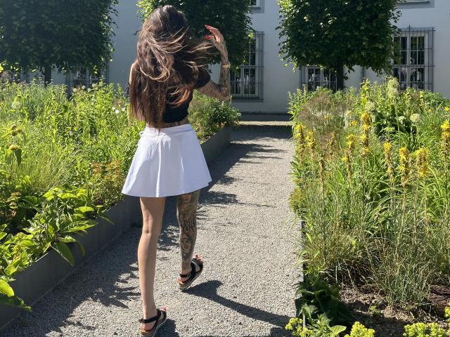 Black-Rosee-hot with long brown hair, black top, white skirt, black sandals, tattooed legs, standing on gravel path, moving right, flinging hair, surrounded by green plants, sunny day.