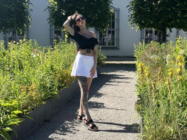 Black-Rosee-hot with tattoos, black top, white skirt, black sandals, standing on gravel path, hand in hair, wearing sunglasses, surrounded by green plants and yellow flowers.