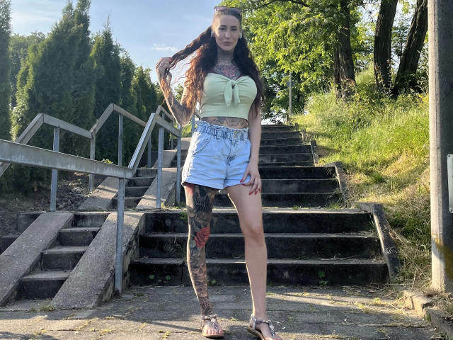 Black-Rosee-hot stands on sunlit stairs wearing a green strap top, blue shorts, and sandals. She has long brown hair, tattoos on her leg, and a confident look.