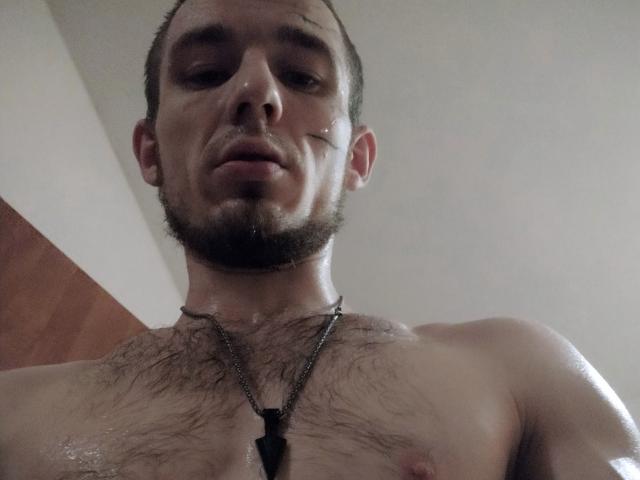 Shirtless FuckPortal with a beard, short brown hair, and chest hair, wearing a dog tag necklace, gazing downward, slightly sweaty, with a serious expression.