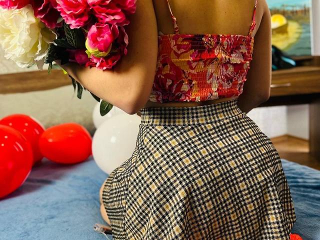 LisaAlisia's back, wearing a colorful floral top and a checkered skirt, holding pink and white roses, with red and white balloons on a blue surface.