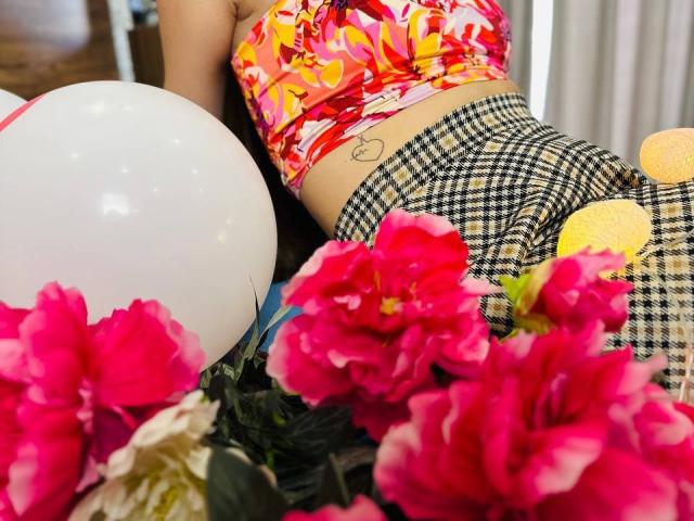 LisaAlisia with pink floral top, black-and-white checkered pants, white balloon, and bright pink flowers. Her ass is tattooed with a heart. Yellow emoji eyes on her butt.