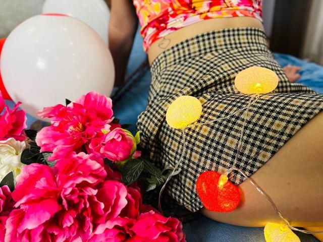 LisaAlisia's ass in checkered shorts, adorned with yellow and orange circular lights, and pink flowers next to a white balloon. Her red floral top is visible.