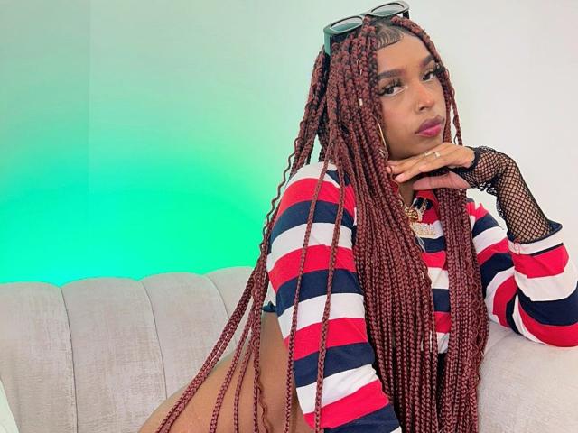 MlleTyra with long red braids, wearing a striped red, white, and blue top, fishnet gloves, and sunglasses on her head, leaning on a white couch, looking serious.