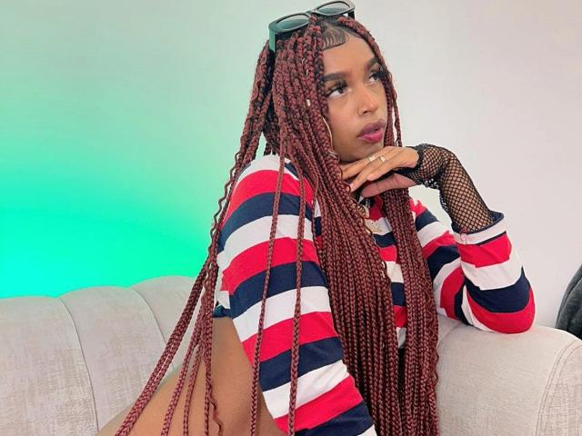 MlleTyra with long red braids, wearing a red, white, and blue striped shirt, fishnet glove, and sunglasses on head, leaning on a white couch with green glow.
