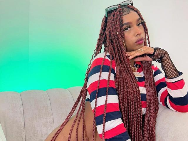 MlleTyra with long red braids, wearing a striped red, white, and blue shirt, fishnet sleeves, and glasses on her head, resting her chin on her hand.