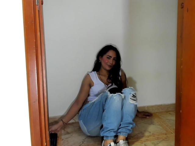 AfroditaHan with long dark hair, wearing a white tank top and ripped blue jeans, sitting on a tiled floor in a doorway, smiling slightly, with her arms behind her.