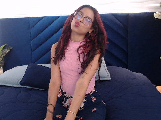 RouseGilmore with wavy red hair, pink sleeveless top, floral pants, pouty lips, glasses, tattoos, bracelet, sitting on dark blue bed with pillow.