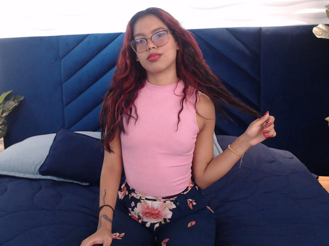 RouseGilmore, con capelli rossi, occhiali, top rosa senza maniche, pantaloni floreali, seduta su un letto blu, che si tiene i capelli, unghie rosse, braccialetto, tatuaggio sul braccio destro, con un leggero sorriso..