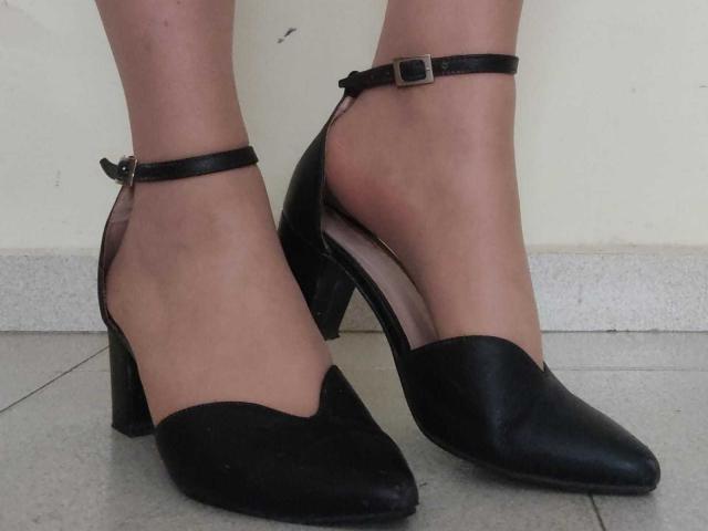 JackyEmma's feet in black leather, pointy-toed, high-heeled shoes with ankle straps and small buckles, standing on gray tiled floor. Shoes have chunky, black, wedge heels.