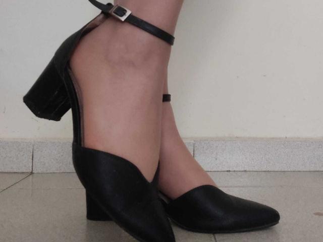 JackyEmma's black high-heeled shoes with a strap, showing her bare legs and feet on a tiled floor. The heels are chunky and her skin is light.