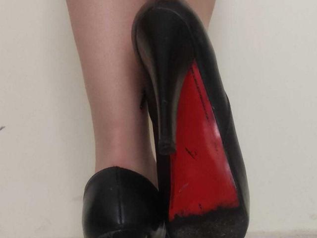 JackyEmma's black high heels with red soles are stacked, showing the worn underside. The heels are shiny with visible scuff marks and her legs are slightly apart.