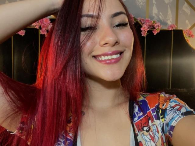 ValeryHarllow with red hair, smiling with eyes closed, wearing a colorful floral shirt, has her hand in her hair, showing her white teeth and pink lipstick.